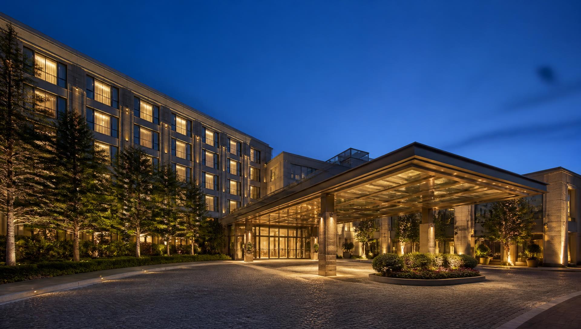 Exterior of a full-service hotel at dusk with lit guest-room windows and a covered entry drive